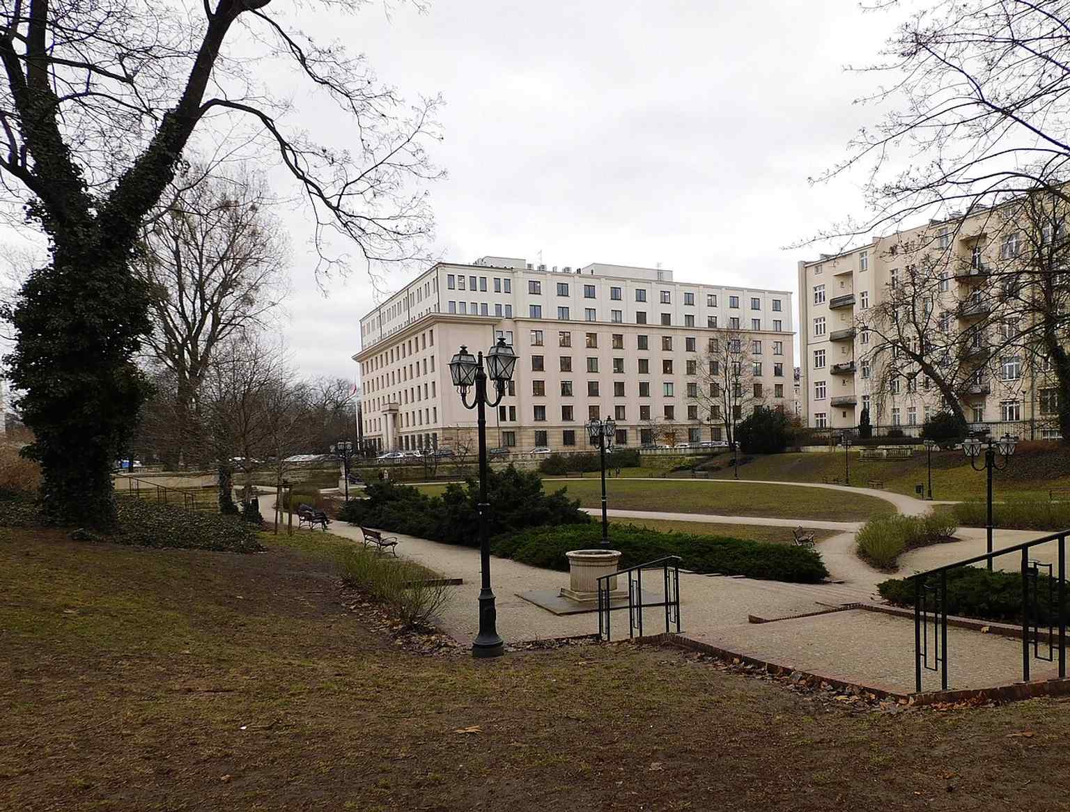 A current view of Swiss Valley with the former PZPR headquarters behind it, showing how the surviving garden fragment now sits against later government-era architecture.
