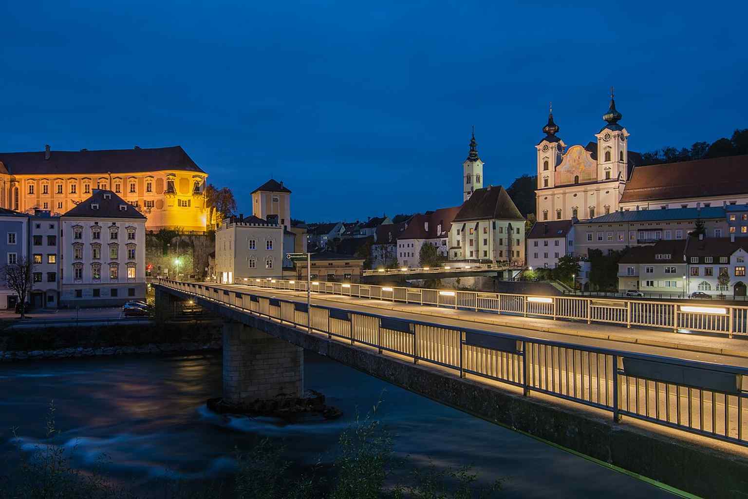 The church in relation to the Enns bridge and nearby historic buildings, highlighting its urban setting beside the river confluence.