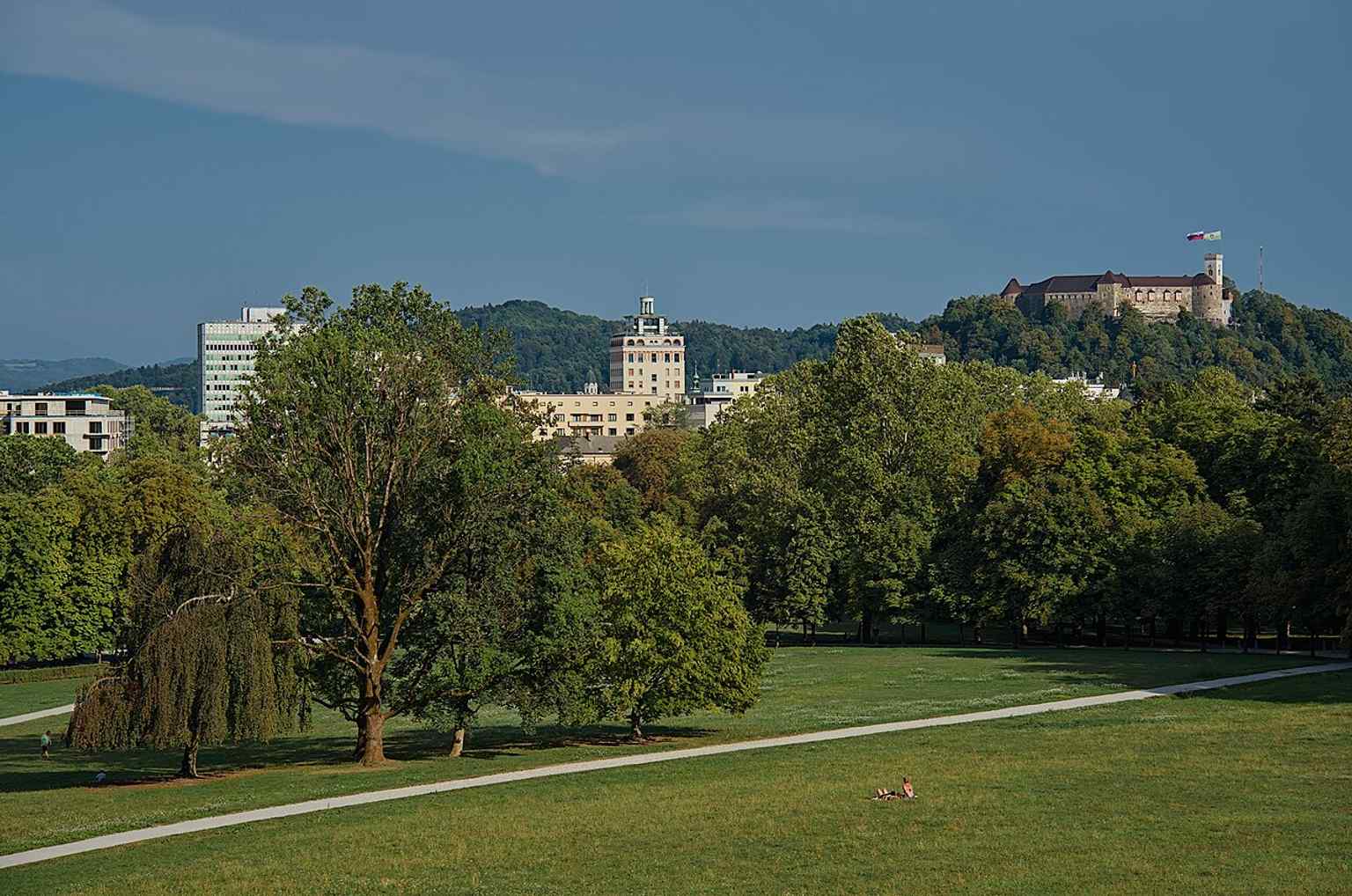 An expansive summer view of Tivoli City Park, a protected nature park since 1984, with Ljubljana Castle visible in the background.