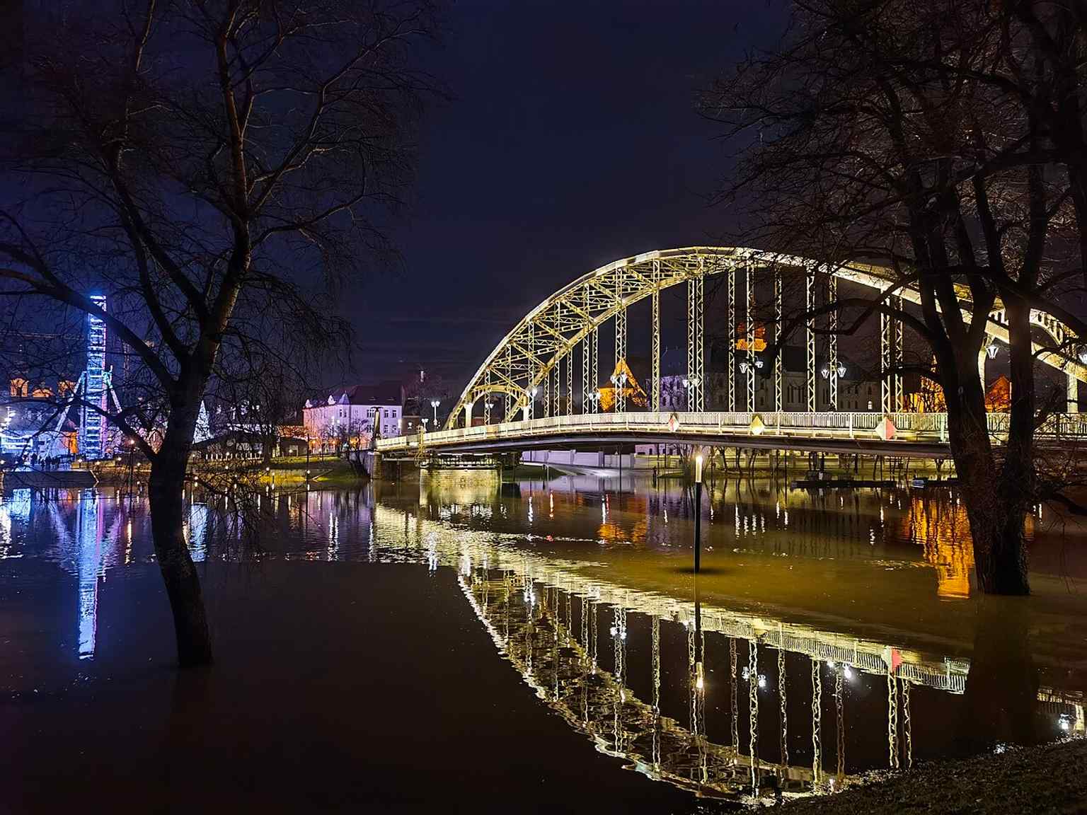 A night-time reflection shot that emphasizes the bridge’s lighting and its strong visual presence over the Mosoni-Duna.