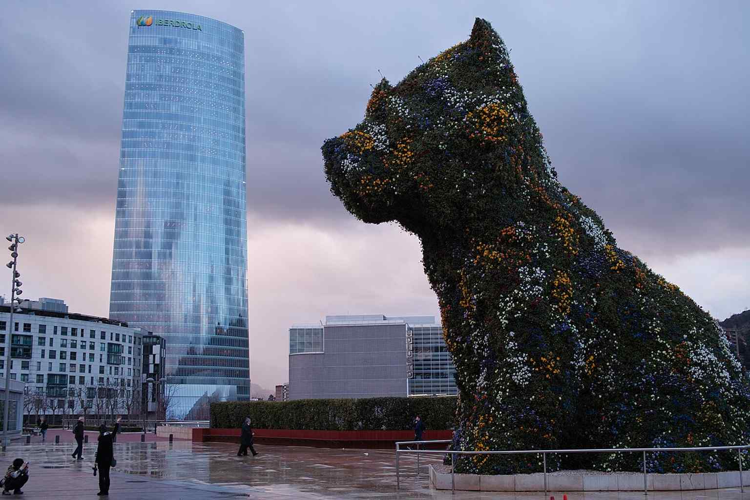 A wider cityscape view of Bilbao with Puppy and the skyline, helping place the sculpture within the modern riverfront setting.