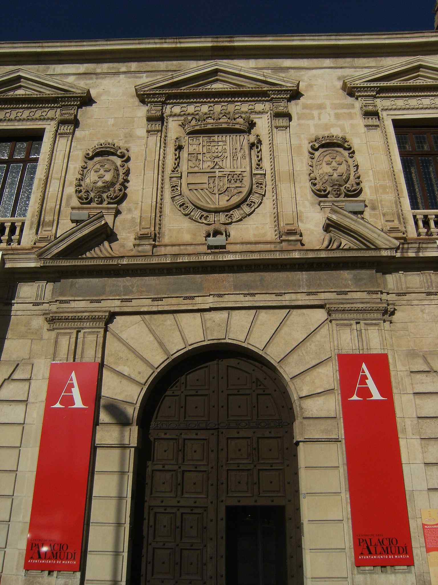 The main entrance features a semicircular arch and a broken pediment displaying the royal Habsburg shield, flanked by two Murcia shields with 6 crowns, predating the reign of Philip V.