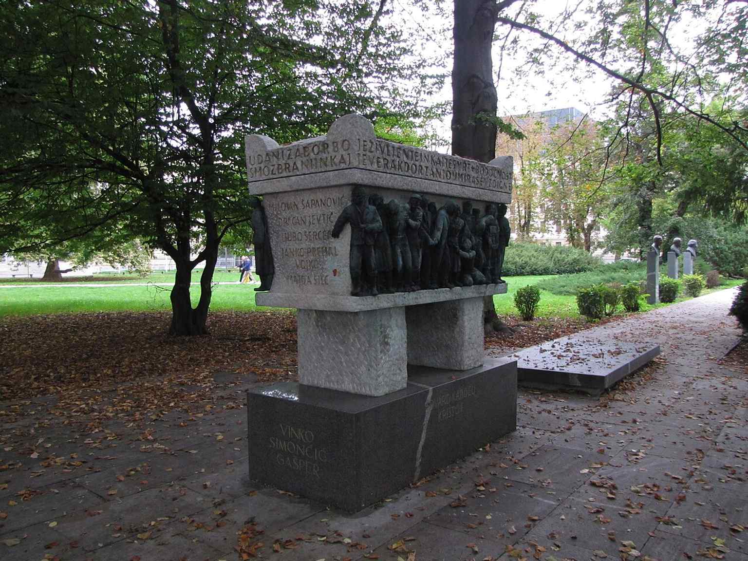 This image captures the stone sarcophagus of the Tomb of National Heroes, designed by architect Edo Mihevc and sculptor Boris Kalin in 1949. Its eastern and western faces are covered by bronze reliefs depicting scenes from the Second World War, and it features a patriotic epitaph by poet Oton Župančič along its top edge.