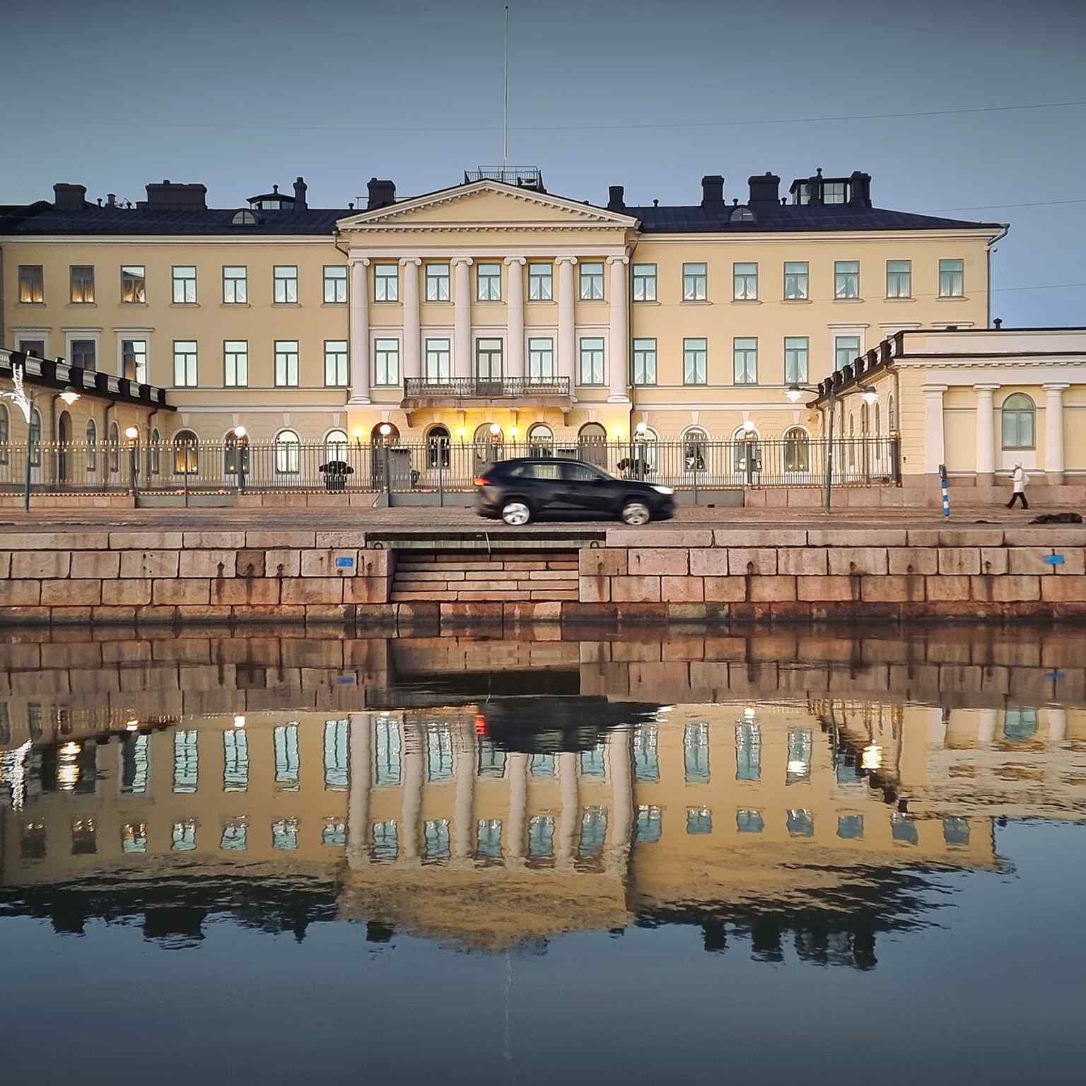 A reflective 2023 view that captures the palace’s façade from an atmospheric angle, modern Helsinki mirrored in the water.
