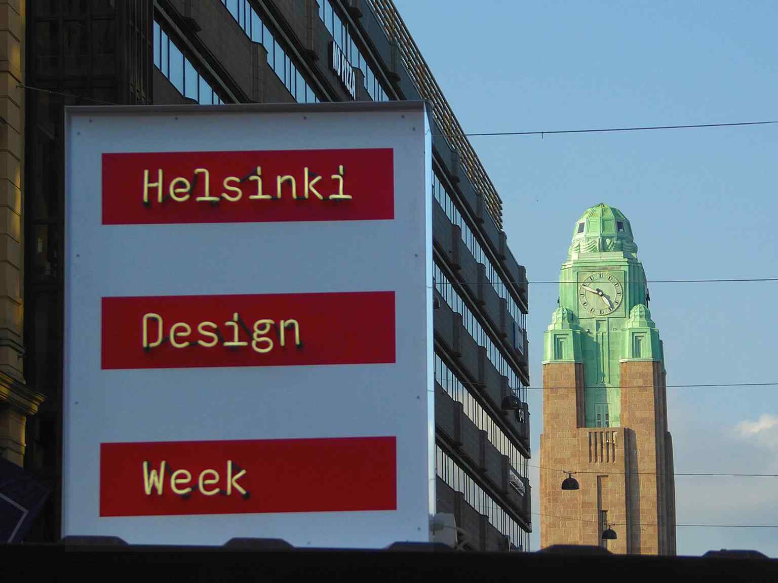 Makkaratalo seen beside Keskuskatu's neon signs and the railway station clock tower, showing how the mall sits in Helsinki's busiest city-centre block.