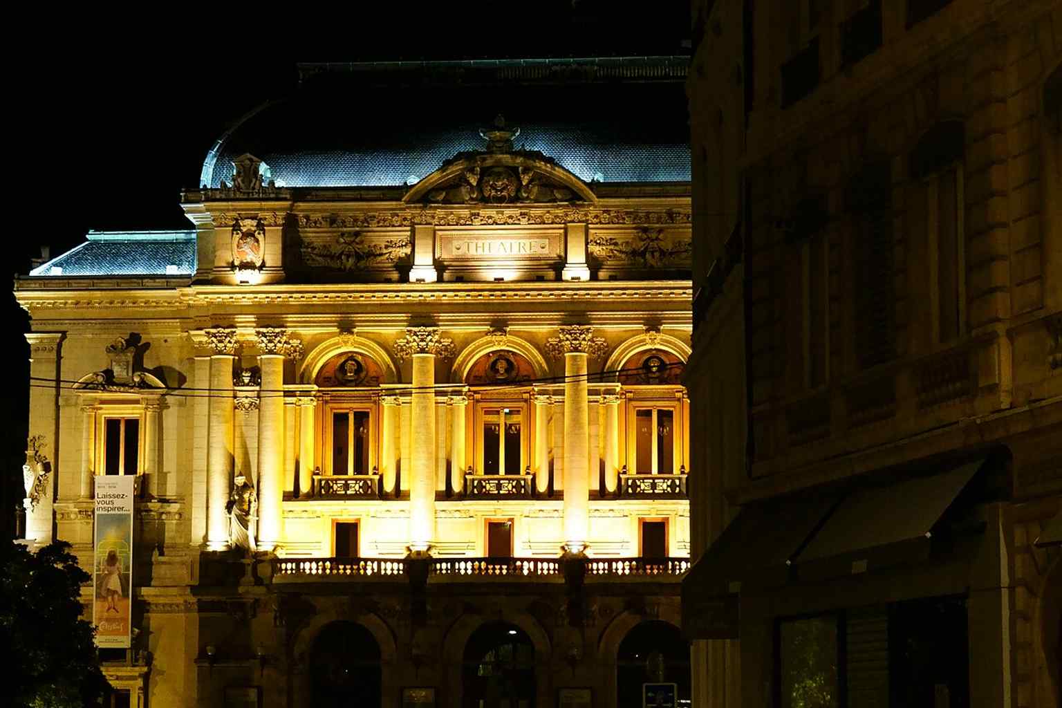 A night view from Rue des Archers, showing how the theatre becomes a lit landmark after dark.
