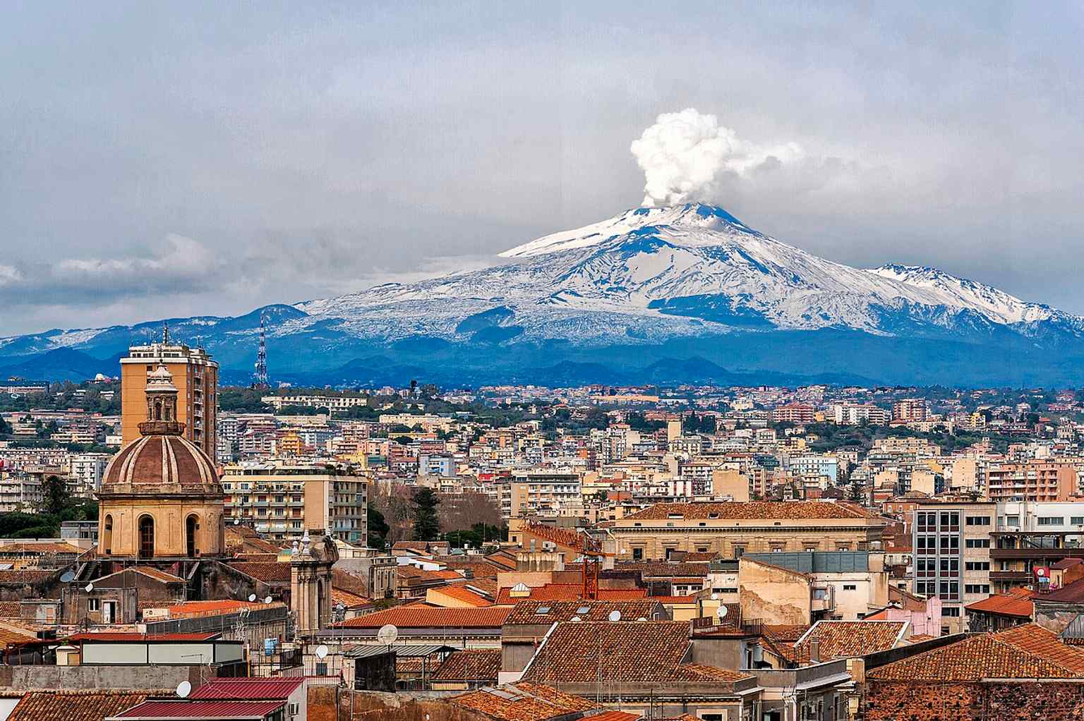 An elevated panorama over the historic center with Mount Etna in the background, echoing how the city’s landscape is shaped by volcanic history.